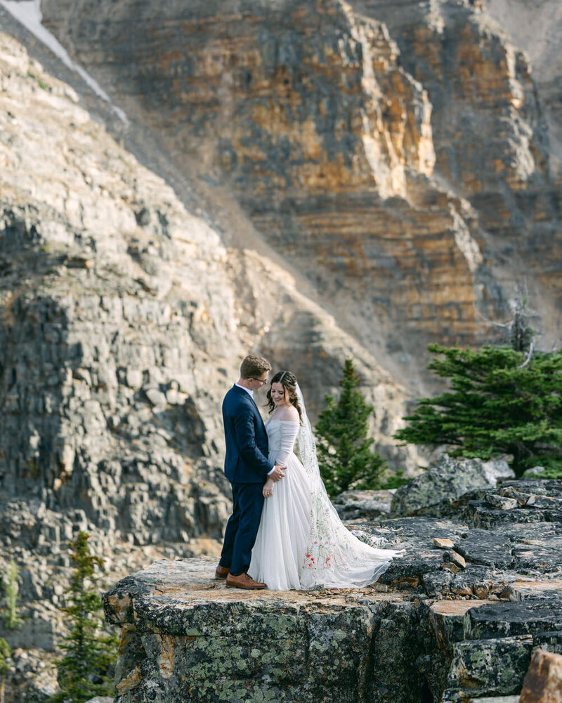 This breathtaking portrait captures the bride and groom holding each other at the top of their adventure hiking elopement at Moraine Lake's Tower of Babel in Banff National Park. The dramatic mountain backdrop adds to the sense of adventure and romance on their unforgettable day.