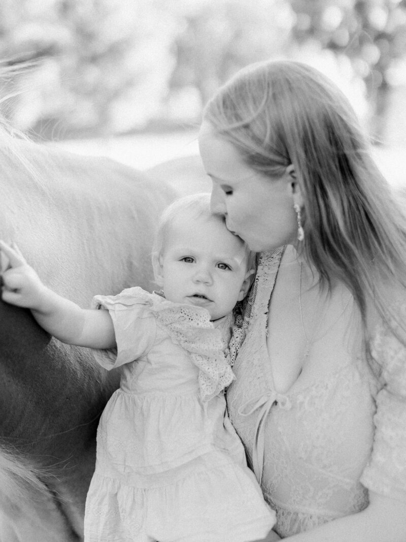 Black and white photo of a mom holder her baby and kissing her head while baby pets their palomino horse on a horse farm by NH family photographer Fieldstone Studio.
