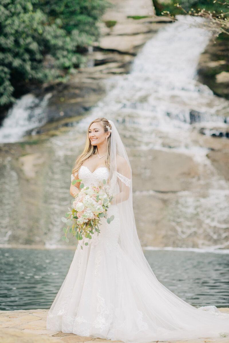 Bride standing gracefully before a cascading waterfall, radiating timeless beauty and confidence on her wedding day. A serene and romantic bridal moment captured in nature