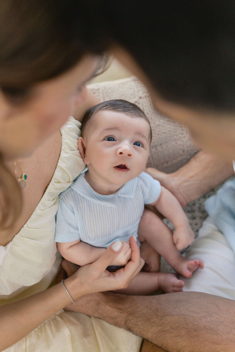 A newborn baby in a blue outfit gazes upward while being lovingly held by both parents, their faces softly framing the moment from above.