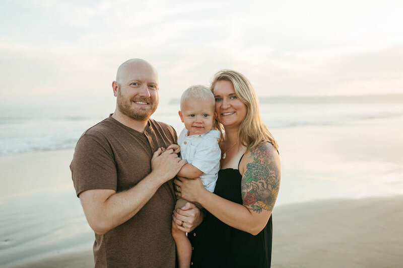 Molly, a San Diego photographer, standing on the beach with her partner and their baby, smiling at the camera during a family portrait session