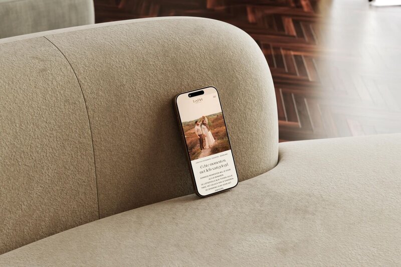 Smartphone leaning against the back of a light beige sofa, screen displaying a photography website homepage featuring a couple in a golden field at sunset.