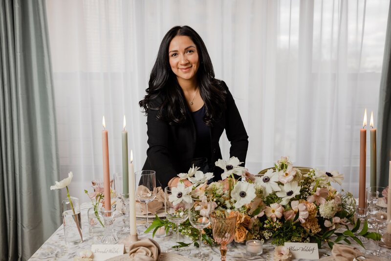 Kathiya, owner of Zela Events smiling while styling an elegantly set table with white florals, candles, and glassware.