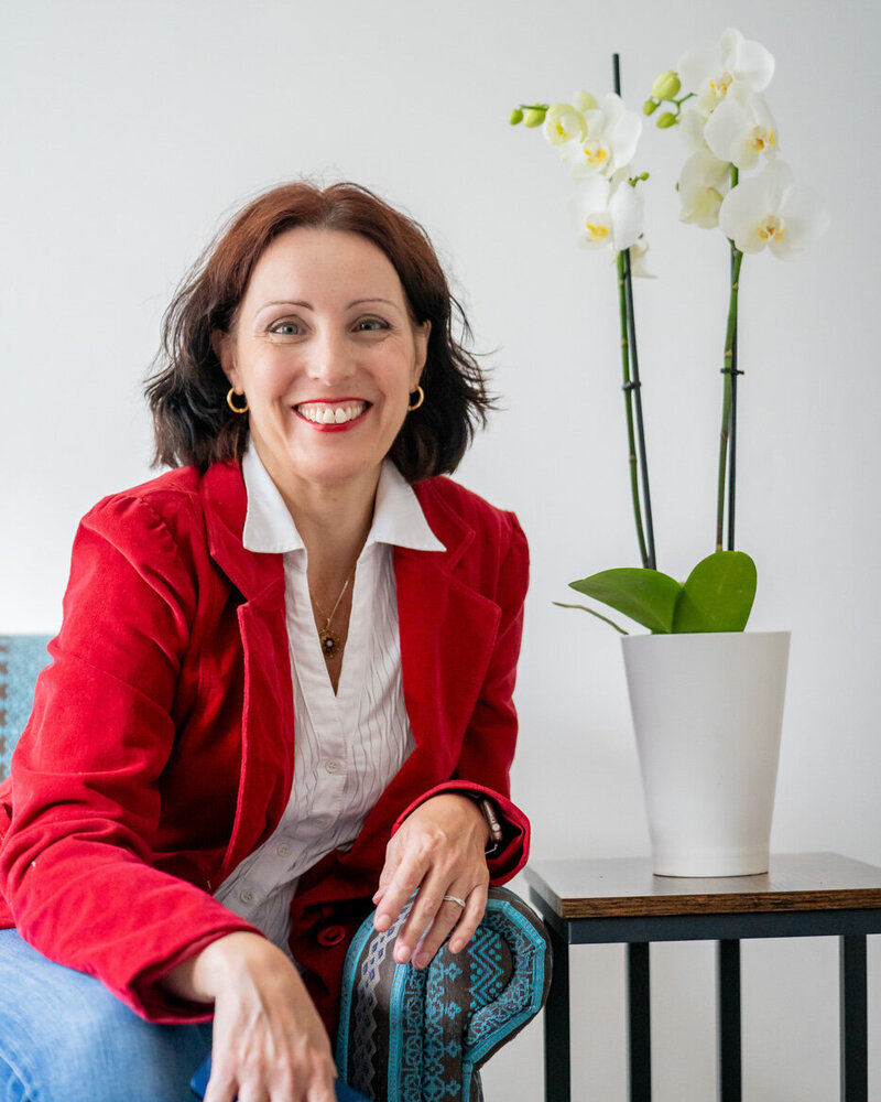 Csilla smiling directly into the camera, leaning slightly forward and leaning to the side of the armchair.