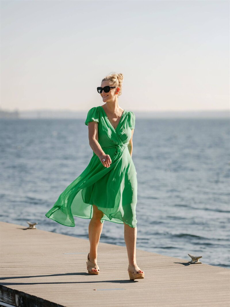 Phillipa Leseberg in a green dress and sunglasses strolls along a wooden pier near a body of water, her hair in an updo like an author deep in thought, and wearing wedge sandals as if she's stepping straight out of the pages of a memoir.