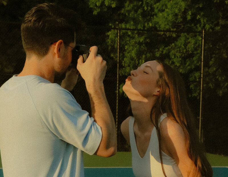 A man is taking a picture of his wife with a film camera. She is posing for him and kissing the air.