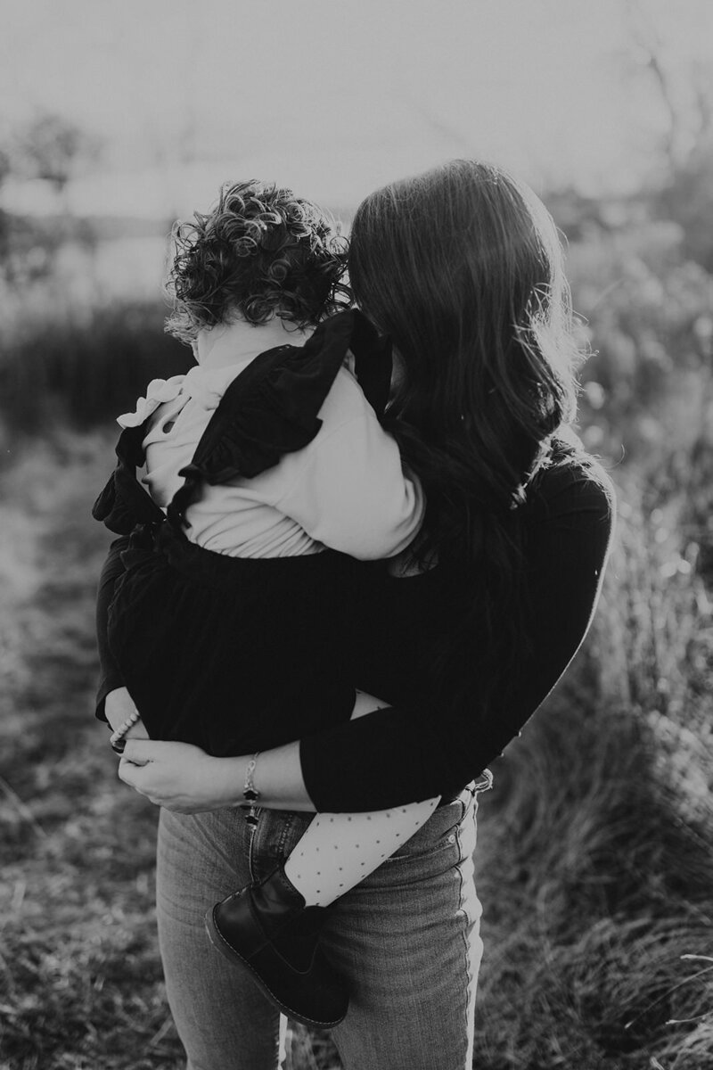 Black and white image of mom cuddling toddler, while toddler nuzzles into mom's shoulder with a sun haze shining over them.