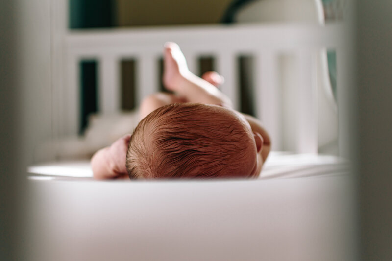 A baby is laying on a pink sheet in a white crib. The photo shows the top of the babies head and feet peeking up in the background.