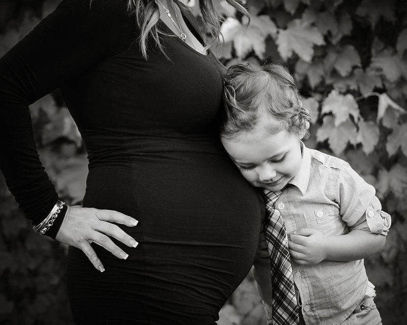 Sky 9 Studio | Little boy leaning on mother's pregnant belly with his eyes closed; black and white photo in front of ivy covered wall