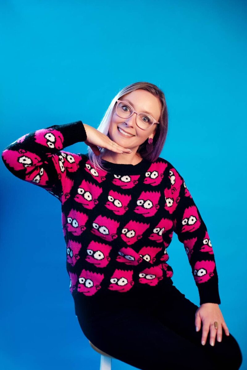 Crystal Genes Photography makes a funny pose while sitting in front of a bright blue backdrop wearing a sweater covered in pink bart simpson heads