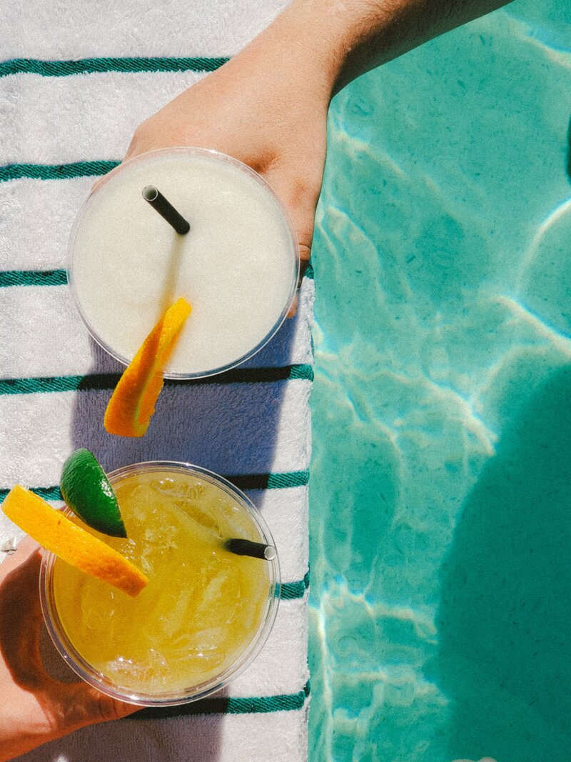 one frozen and one chilled margarita being held by two hands at the edge of a pool for the photography services page socialfizz