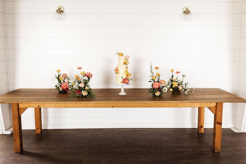 A large wooden farmhouse table holds a three tier wedding cake on a white pedestal cake stand. The cake sits in the middle of the table, surrounded with little arrangements of fresh flowers. The cake is pale yellow, and decorated with the same types of flowers on the table around it - in pinks, yellows, oranges.