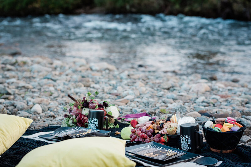 A beautiful luxury charcuterie and fruit platter with a basket of colorful macaroon cookies sits in a beautiful styled elopement picnic on the rocky shore of a river.  This picnic is provided for the eloping couple during their Alaska Glacier Elopement package in Denali National Park.  The brides moody colored floral bouquet rests on the picnic blanket as well. 