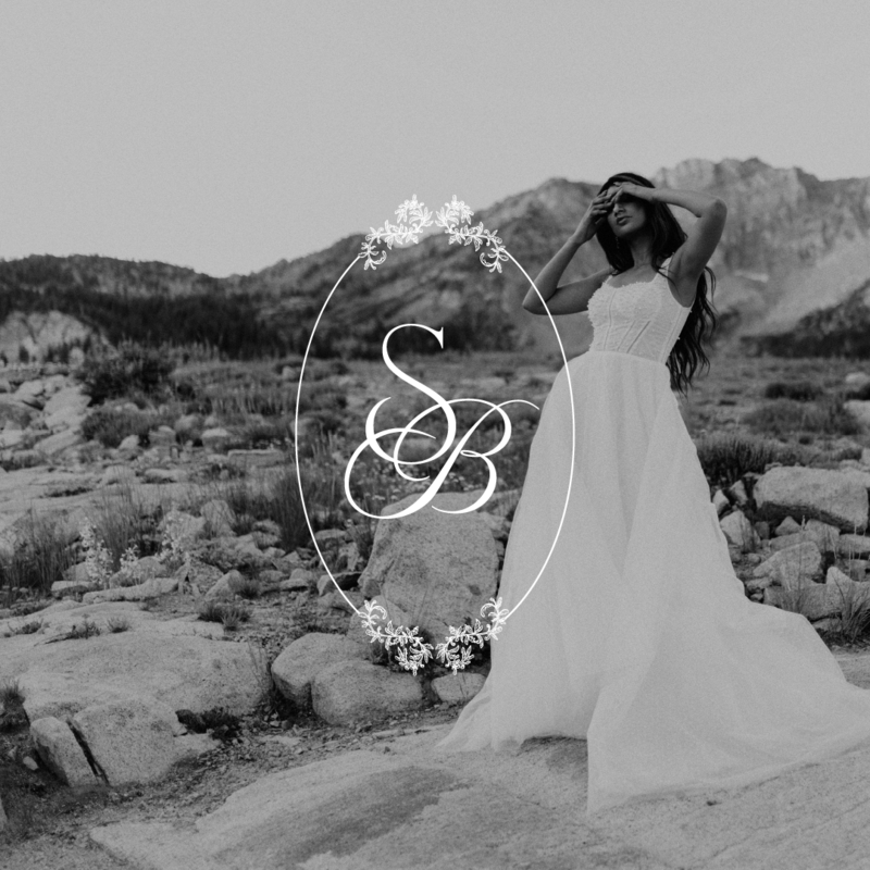 A women in a flowy wedding dress is standing in front of a rocky mountain, covering her face, while there's an overlay of a bridal salon's logo over the photo. The logo has the letters "SB" and is incased by an oval with floral features.