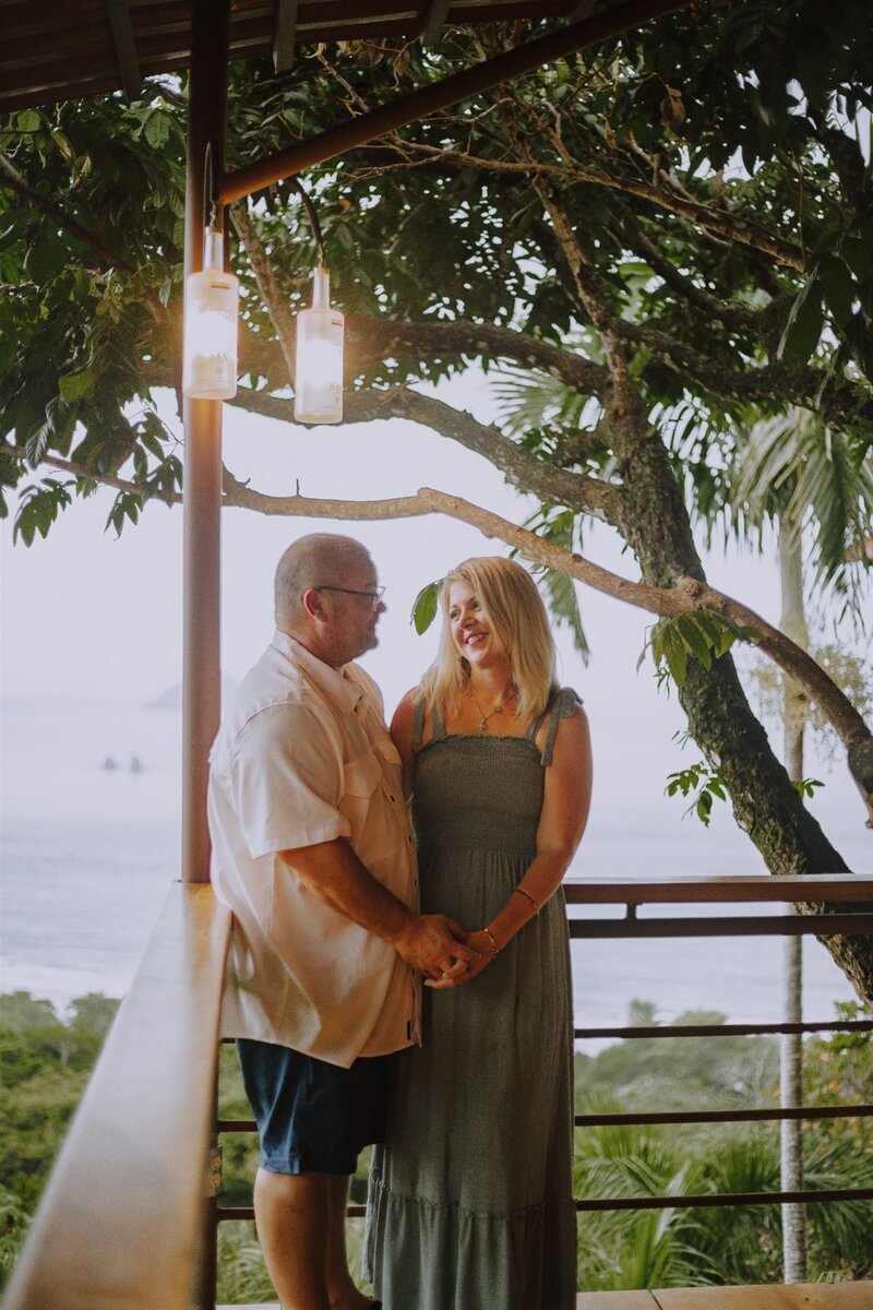 Couple holding hands and smiling on a balcony surrounded by lush tropical trees and ocean views at sunset.