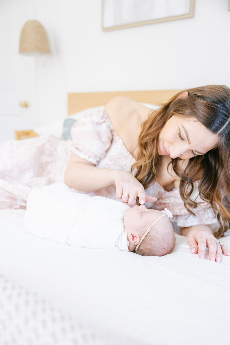 A mother wearing a floral off-the-shoulder dress lovingly gazes at her swaddled newborn during a natural light in-home newborn photography session in Fairfield County, CT, captured by Nicole Neira Photography.