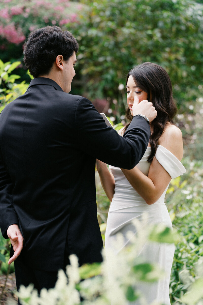 Tender moment between a bride in an off-shoulder gown and groom in a black suit surrounded by greenery.