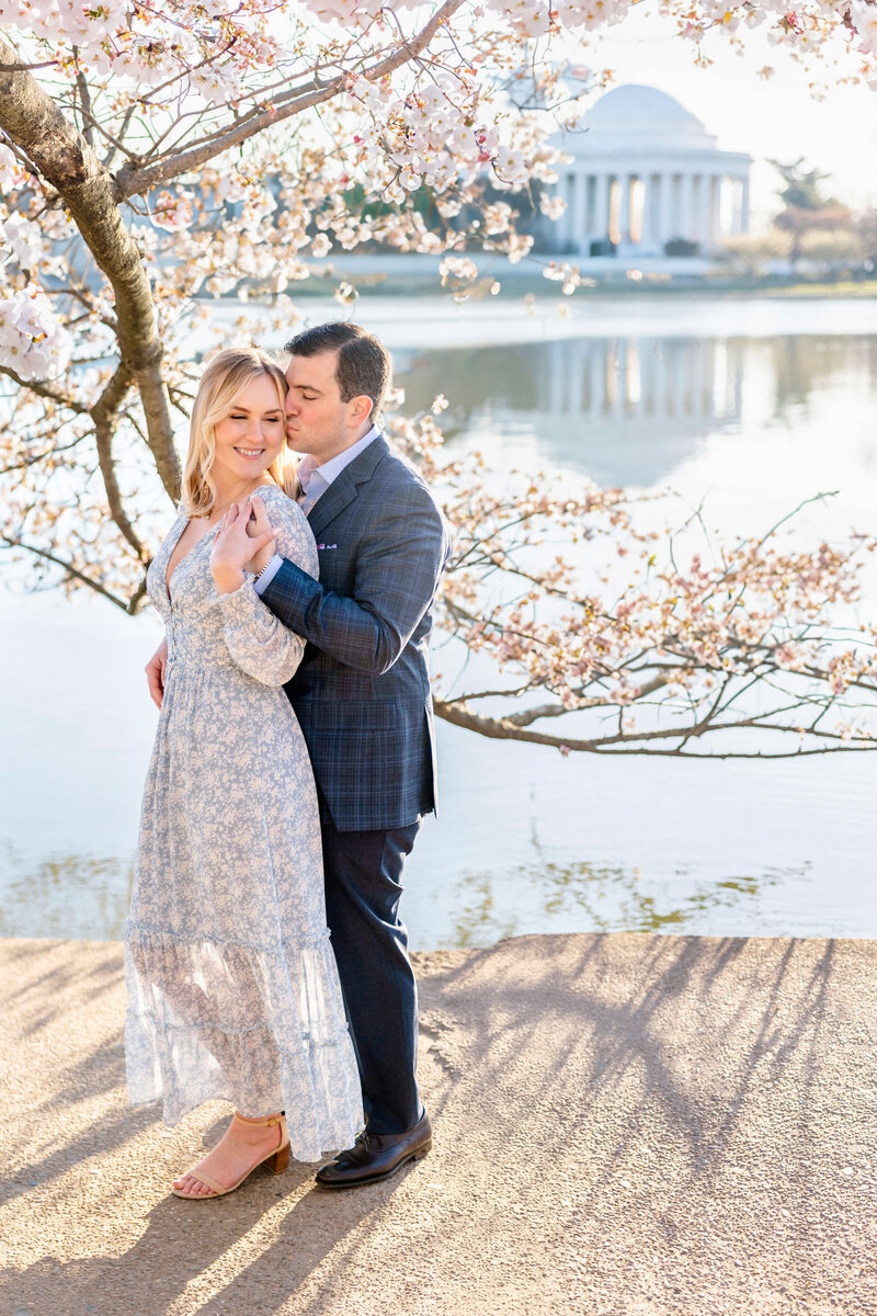 A couple embraces under cherry blossoms by a serene lake, with a domed building in the background. The woman wears a light dress, and the man is in a suit. The sun illuminates the scene, creating a soft, romantic atmosphere.