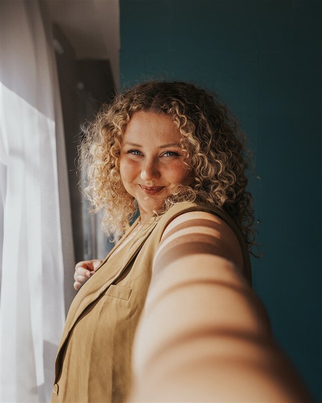 A close-up, self-portrait of Andrea Cakmar smiling into the camera, with sunlight casting shadows on her face. She wears a tan blazer and stands near a window with sheer curtains.