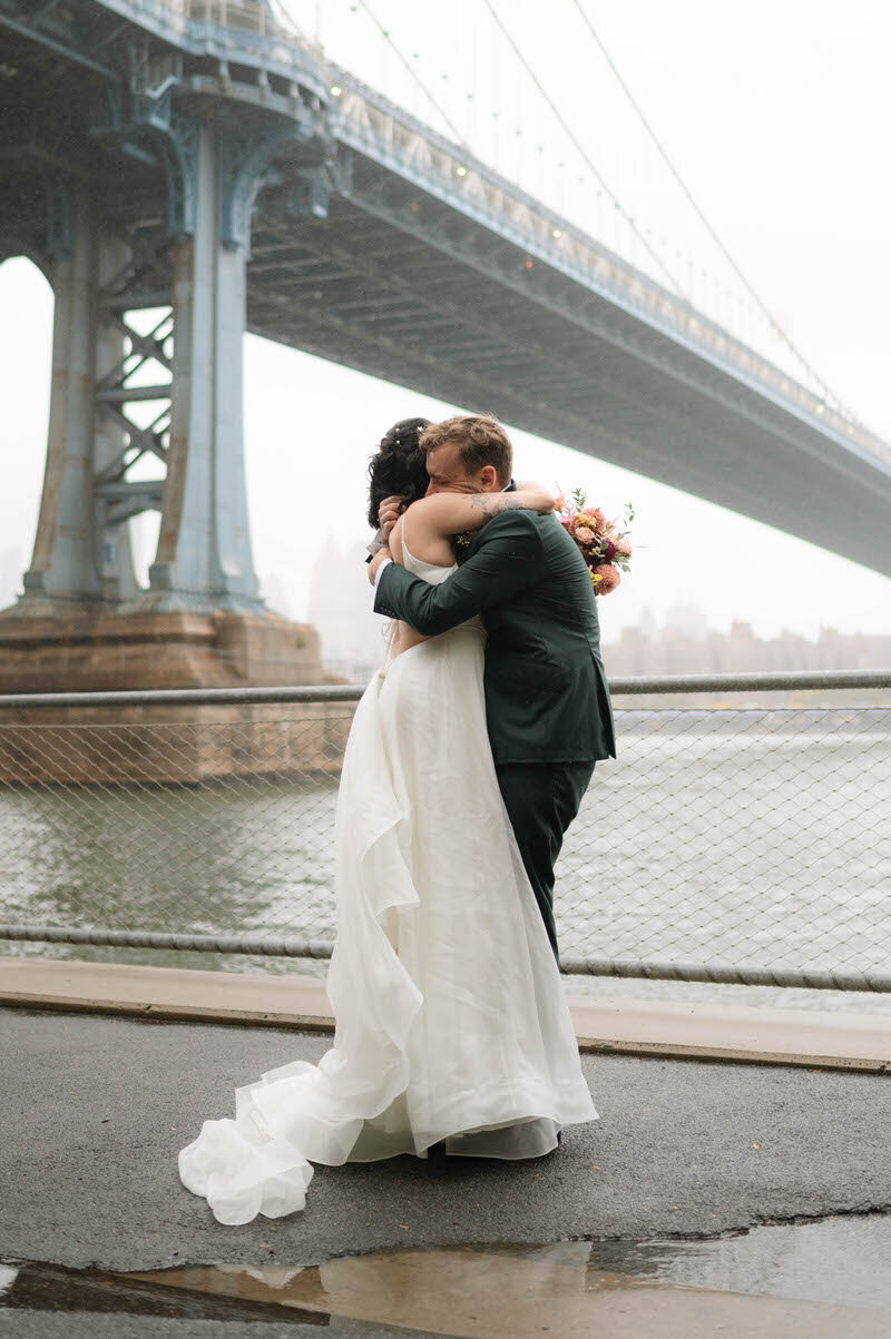 Bride and groom hugging on a dreary day beside the Brooklyn Bridge