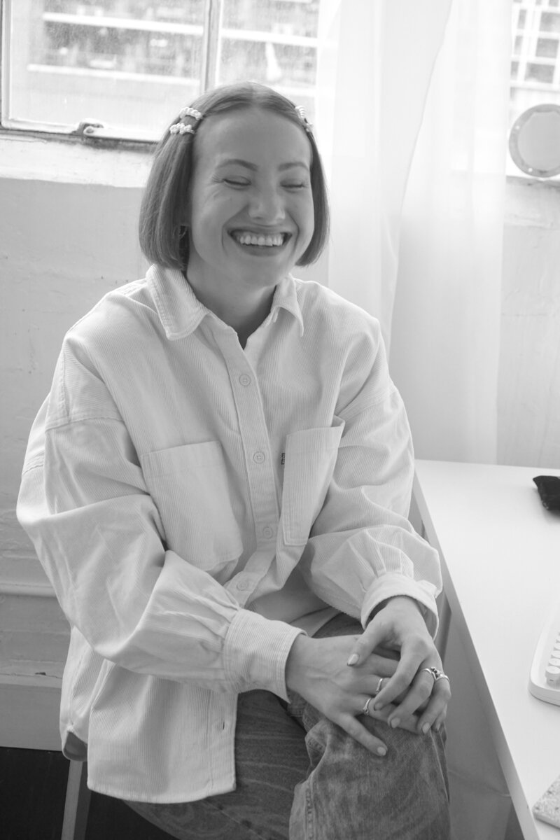 Margarita is sitting on a stool at her desk,  with a curtain in the background; Margarita has a silly grin on her face, with her eyes closed, because she is thinking about all the fun we'll have writing ethical copy together!