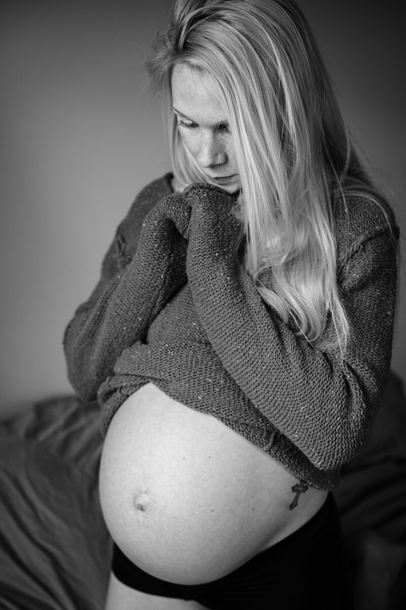 A black and white portrait of a pregnant woman standing with her hands near her face, wearing a cozy sweater and looking down thoughtfully at her bare belly.