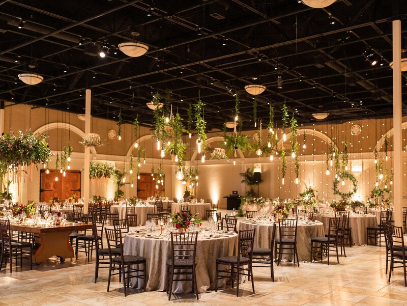 A wedding reception in a spacious room filled with hanging bulbs and greenery.