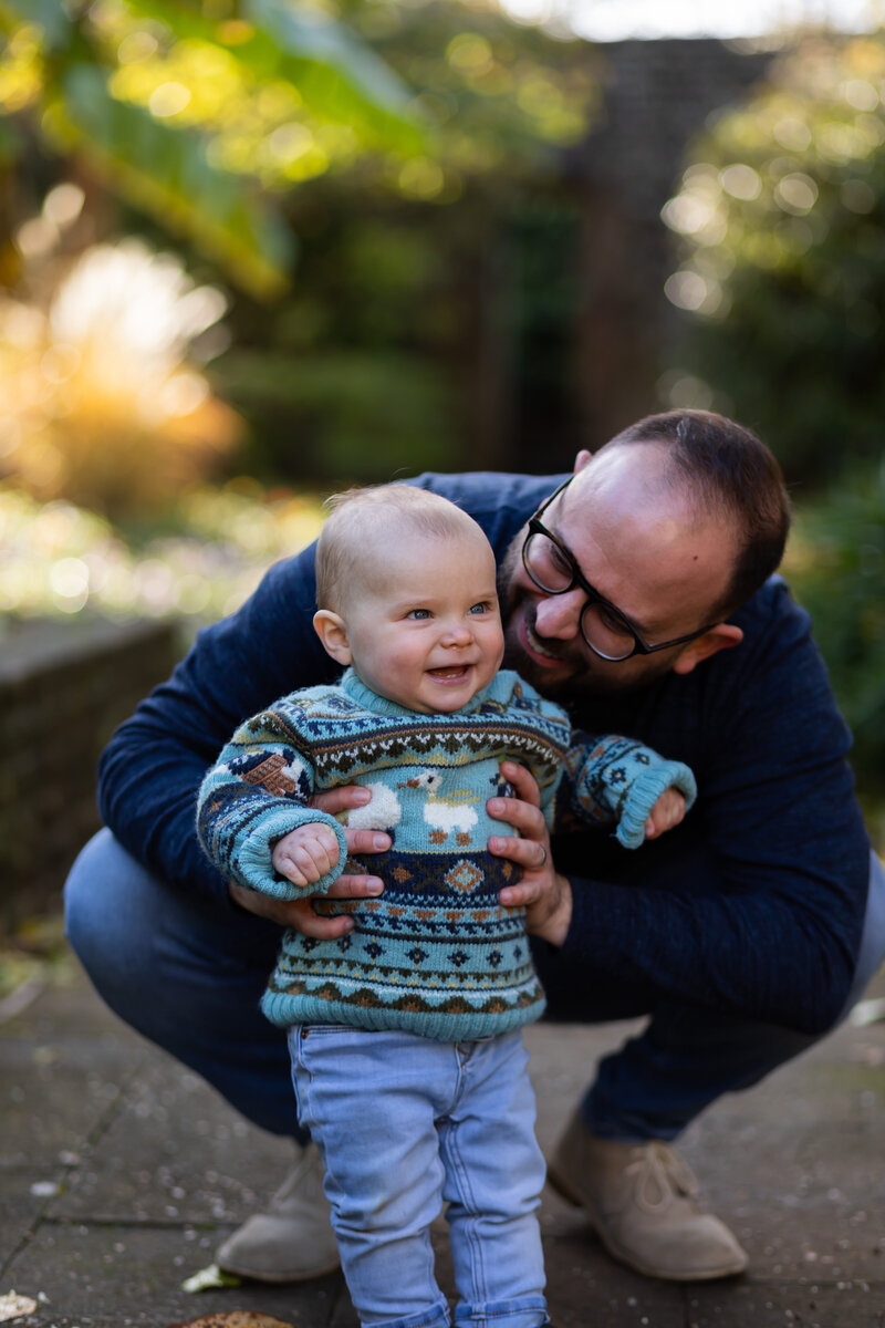 small toddler smiles as dad looks at him from the side holding him up so he can stand