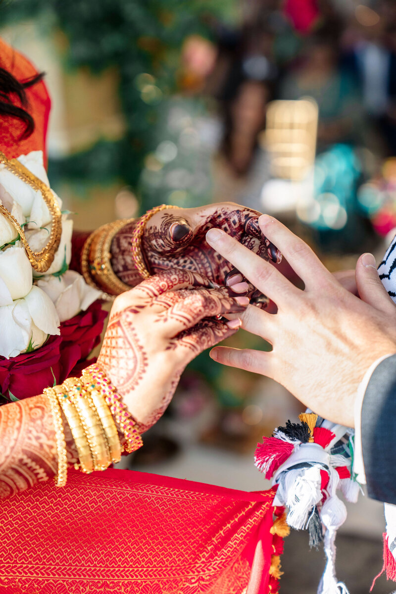 A bride and groom exchange rings during a wedding ceremony. The brides hands are adorned with intricate henna designs and gold bangles, while the grooms hand is being held gently. The scene is vibrant with traditional attire and floral decorations.