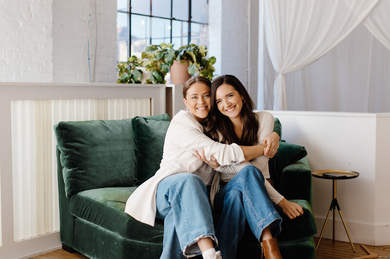 2 women hugging on green couch with plants in background