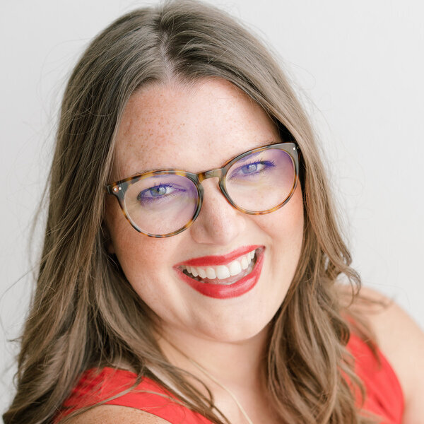 A woman with long, wavy brown hair and red lipstick smiles broadly at the camera. She is wearing tortoiseshell glasses and a red top, and the background is plain white.