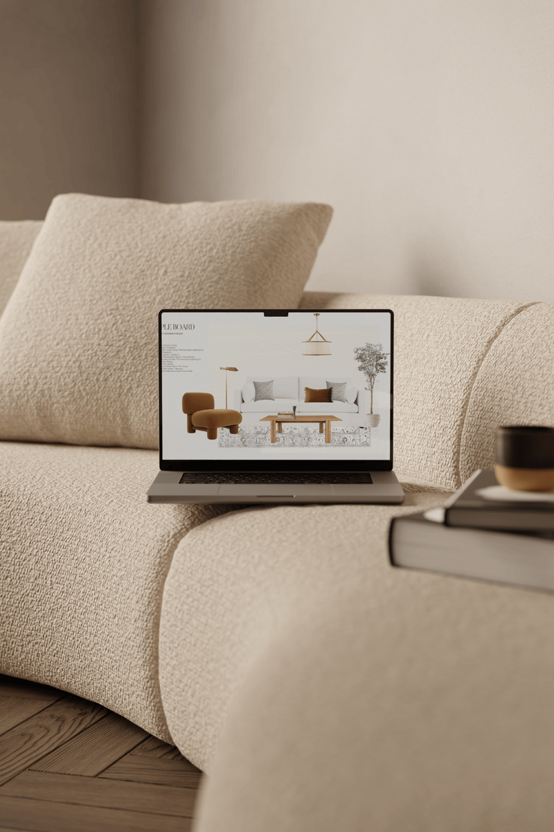 Laptop on a beige bouclé sofa, displaying a sample board for a living room design, with books and a cup of coffee placed in the foreground on the sofa.