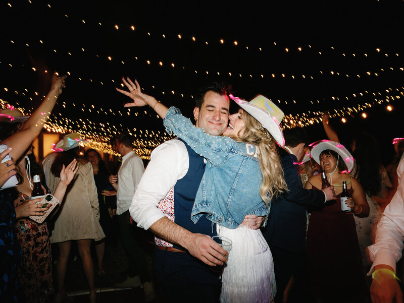 Bride in light up cowboy hat and denim jacket hugging groom on the dance floor under string lights