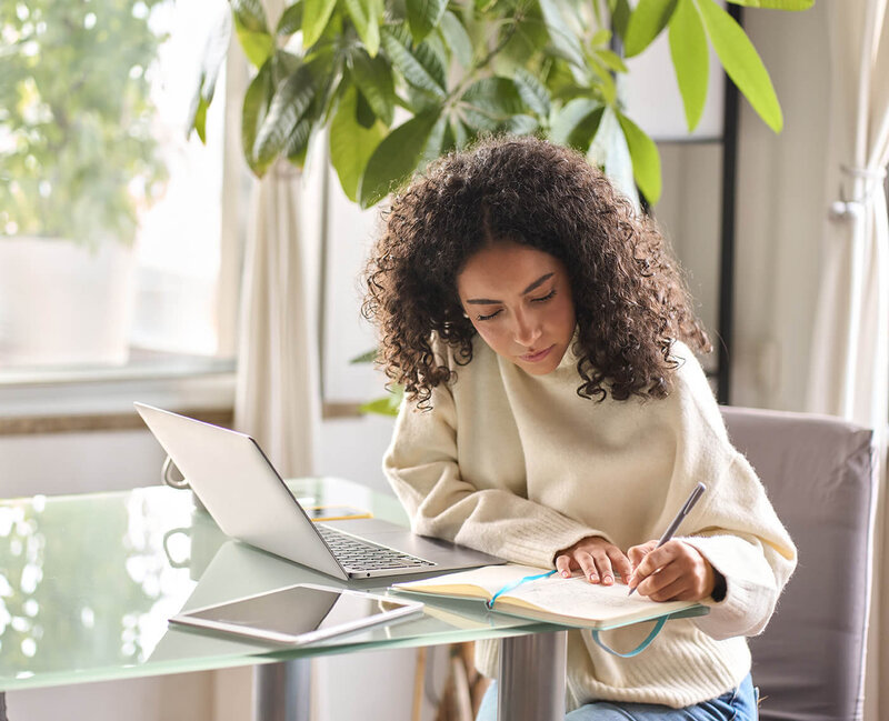 Woman in a cream sweater  sitting at her desk  with a laptop and writing  in a notebook