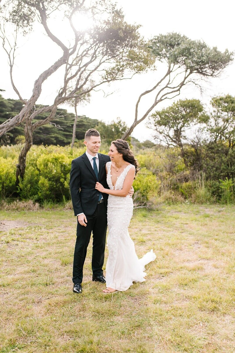 A bride and groom cuddling in an outdoor area under the shade of a tree