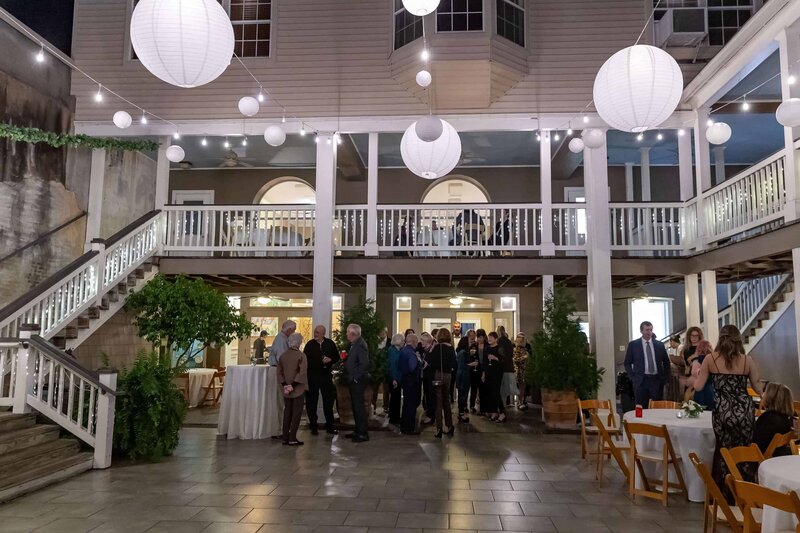 A group of people gathers in a spacious, well-lit courtyard decorated with white paper lanterns and string lights. There are two levels with white railings, scattered tables, and trees adding greenery to the festive scene.