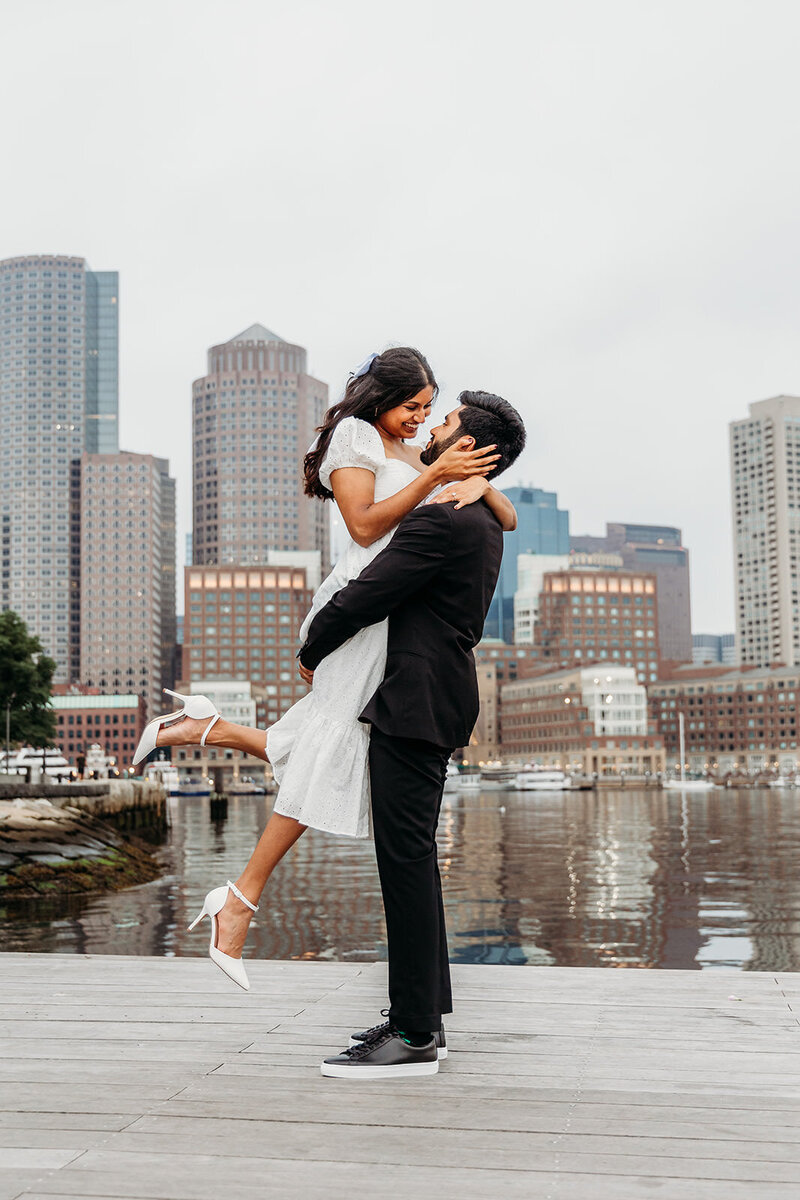 Man lifts his fiance on the Boston waterfront during engagement portraits