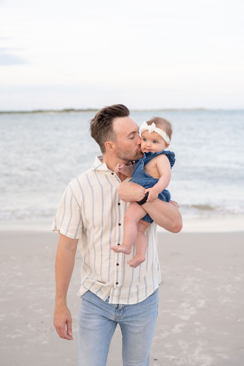 father stands on the beach holding his baby girl with one arm while giving her a kiss on the cheek