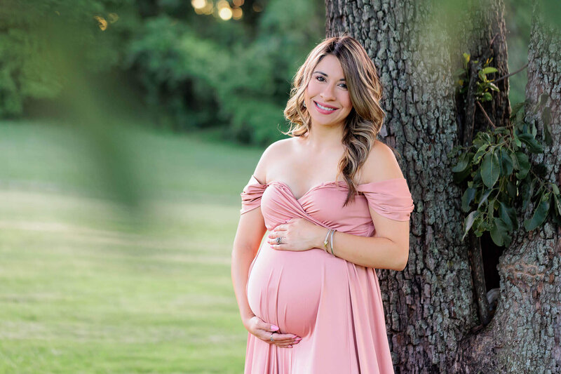 A woman in a pink dress embracing her pregnant belly and leaning against a tree at a park.