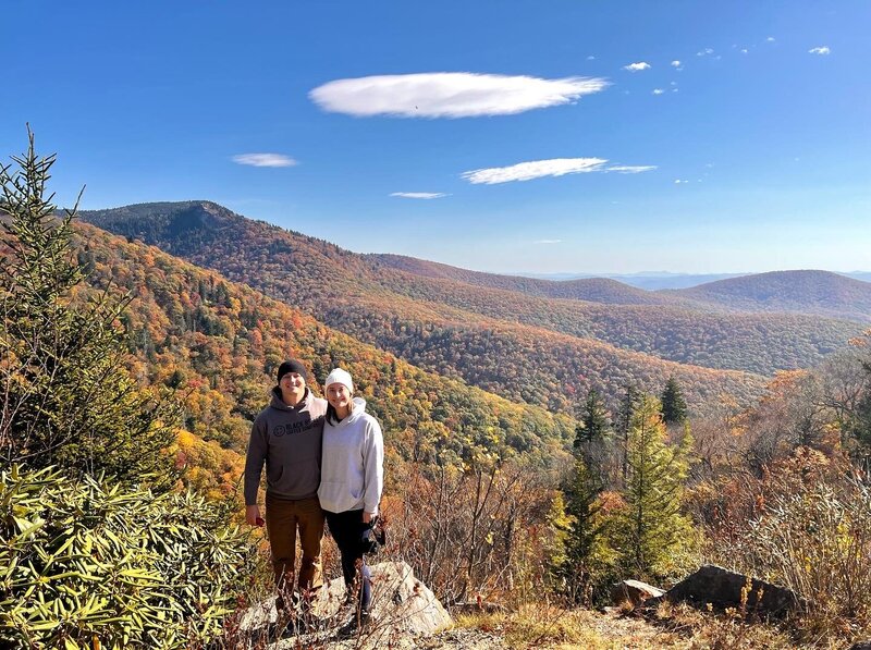 A woman and man stand side by side smiling at the camera in front of a huge mountain range with layers of mountains. The leaves are changing colors turning the mountains shades of yellow, red, orange and gold.