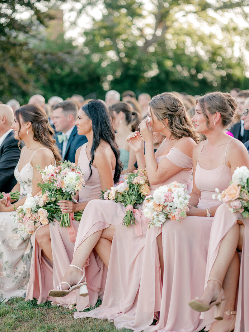Bridemaids in pink sit and watch a wedding ceremony and wipe a tear away from face