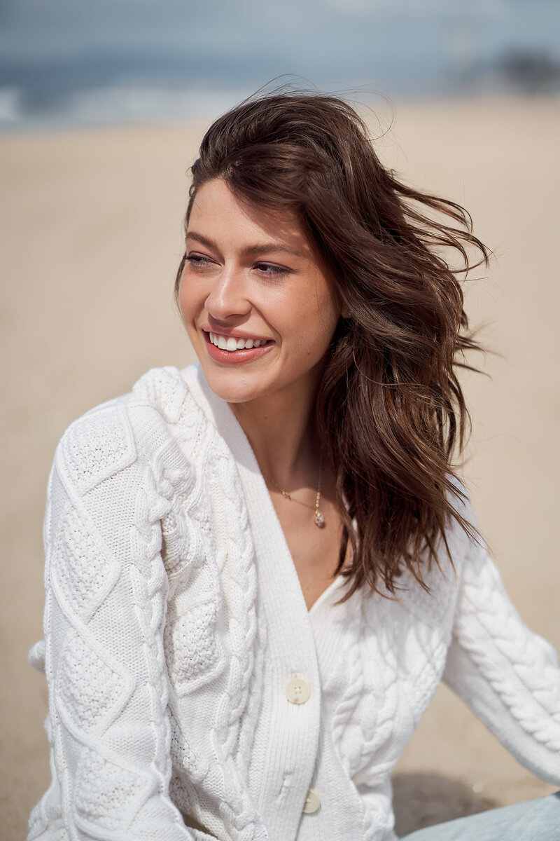 woman smiles big in portrait at the beach wearing a white cardigan with brown hair flowing in the wind