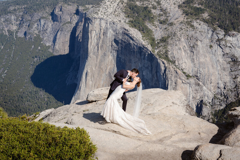 A man dips a woman in a wedding dress and kisses her with El Capitan in the background after saying their vows in Yosemite National Park