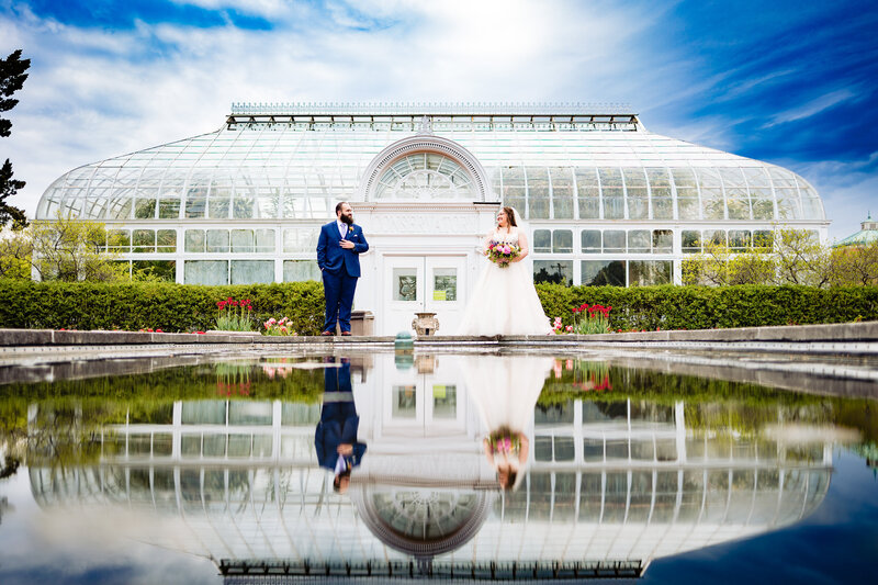 Wedding couple standing apart in front of the conservatory at the Toledo Zoo with the reflection from the water in the foreground
