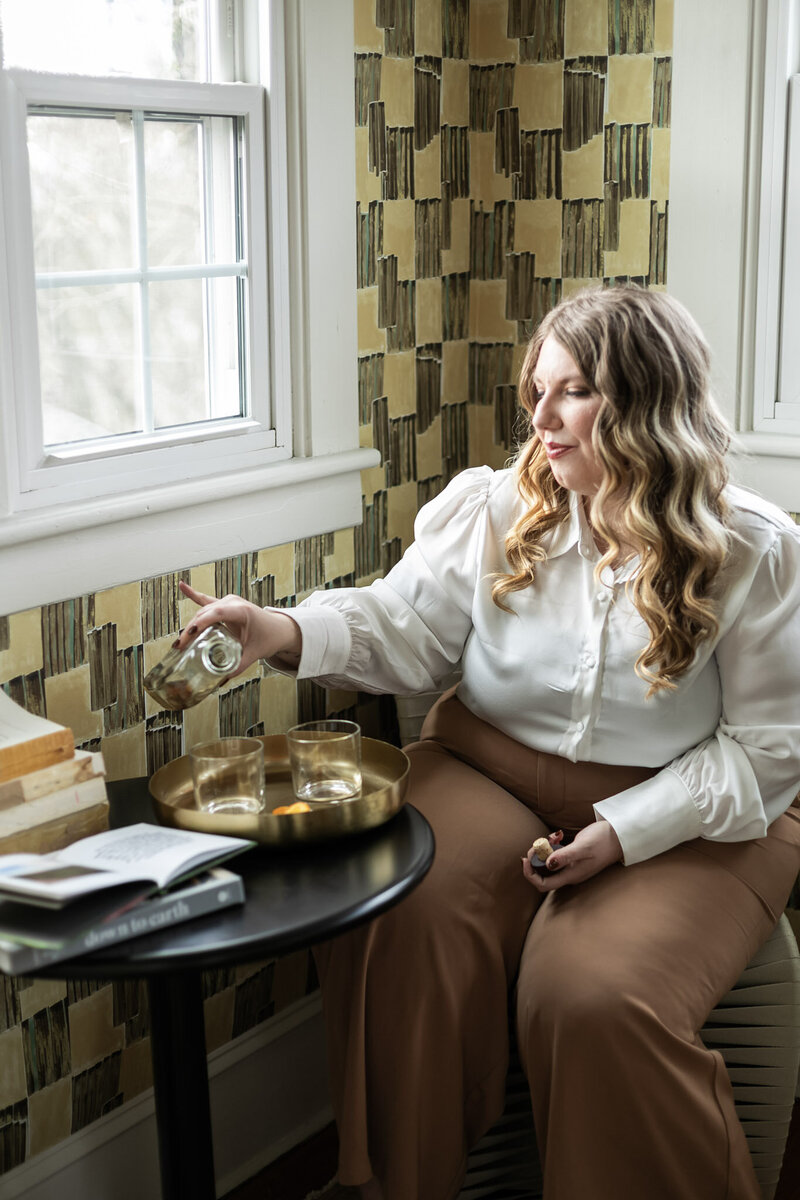 Ashleigh Henry, life coach based in Asheville, North Carolina is pouring an old fashioned in a room at The Blind Tiger, near The Biltmore House and Riverarts Distrct, with green and yellow wall paper behind her, near a window with a small black table holding books and journals. 