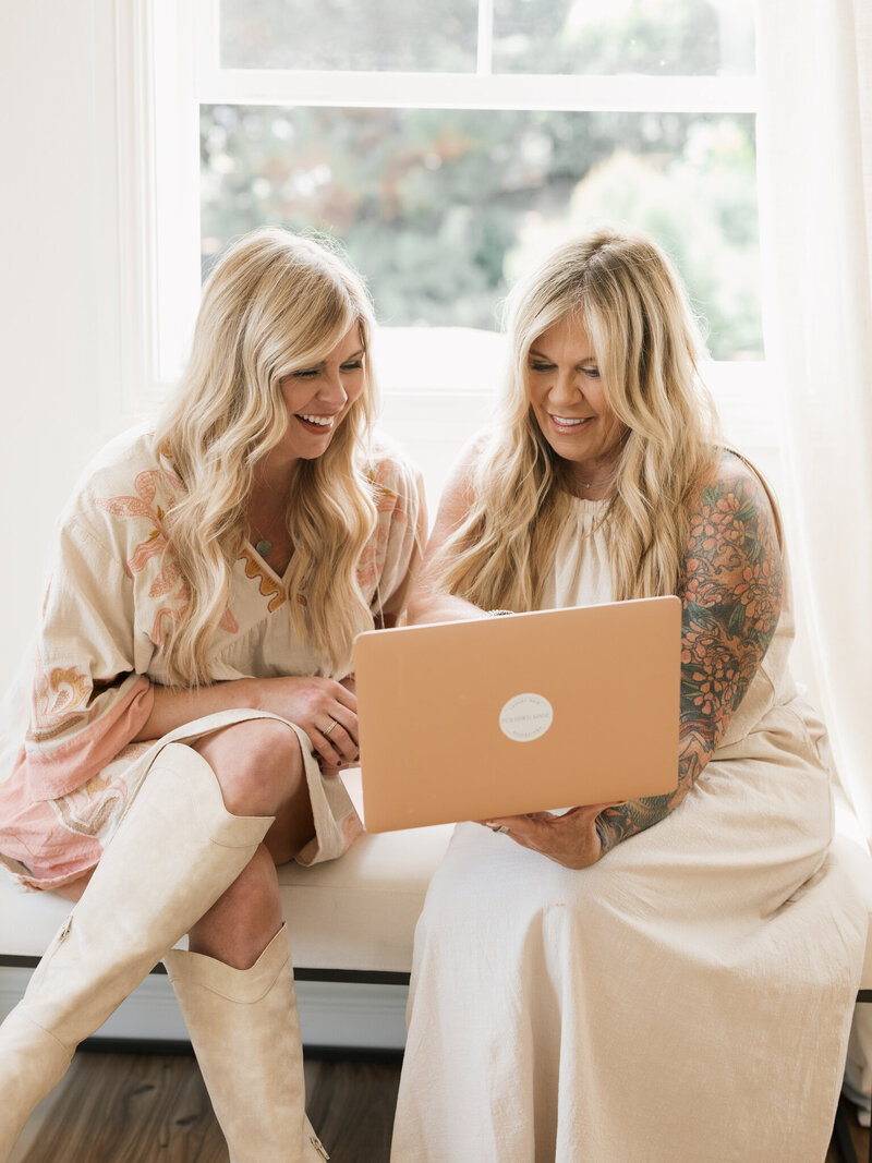 two women sitting in window seat looking at  laptop