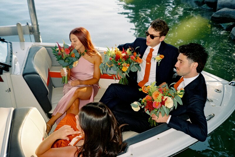 Wedding party members holding colorful bouquets while seated on a boat, dressed in formalwear with an Iowa lake in the background.