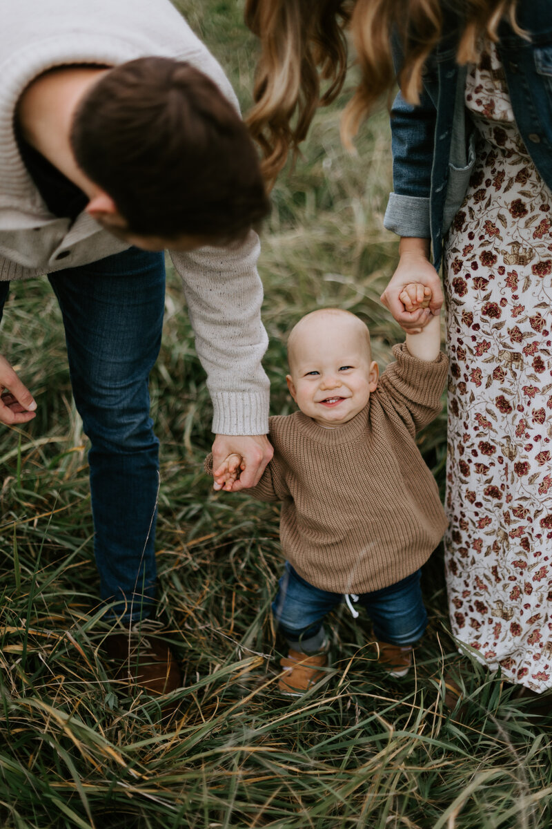 A baby stands in long grasses holding his parents hands and smiles widely at the camera. Family photo session at Prophetstown State Park in West Lafayette, Indiana. 