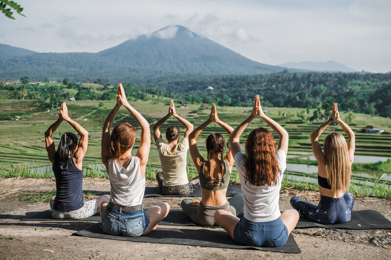 Group of women practicing yoga outdoors with hands in prayer pose, facing a misty volcano landscape — symbolic of wellness, stillness, and transformational travel.

