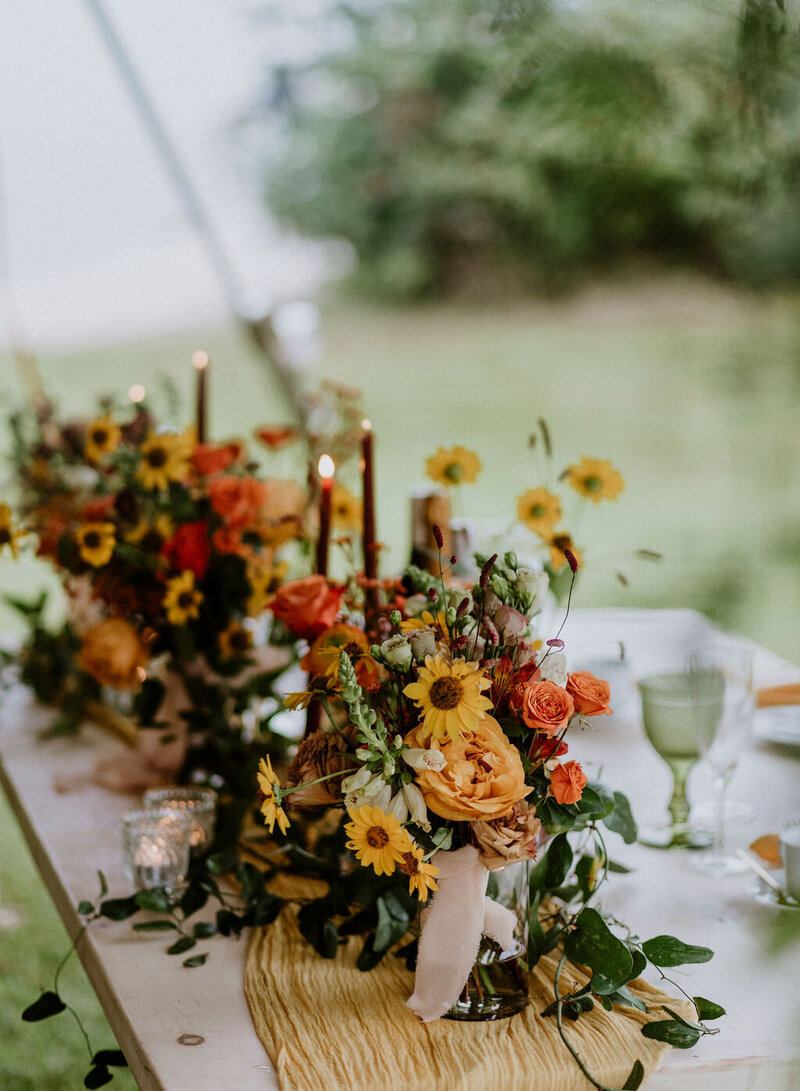 Mustard and burgundy garden flower centrepieces with burgundy tapered candles line a wood harvest table at a private estate wedding under a tent in Cobden Ontario